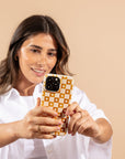 A woman with long brown hair and a white shirt smiles as she takes a selfie against a beige background, holding her smartphone with the Star Terracotta Checker Eco-Friendly Phone Case in yellow and white checks.