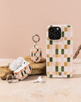 A phone with the Pink and Green Watercolour Eco-Friendly Phone Case, along with AirPods in a matching case and a keychain accessory—all featuring a pastel green, pink, and cream checkered pattern—are displayed on a beige surface with a piece of wood in the background.
