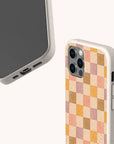 Two phones with beige, biodegradable phone cases—one displaying the front screen, the other showcasing the back of the Pink and Purple Checker Eco-Friendly Phone Case with a pink, orange, and beige checkered pattern. Both eco-friendly cases feature a speckled texture and visible side and bottom ports.