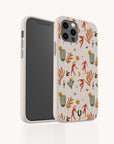 Standing upright against a plain background, the Three Dancers Eco-Friendly Phone Case showcases a beige compostable design adorned with a colorful desert-themed pattern featuring orange and green plants, red animal-like dancer figures, cacti, suns, and abstract shapes.