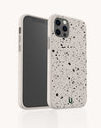 The Black Splatter Eco-Friendly Phone Case in beige features a minimal design with black splatter patterns, triple rear camera cutouts, and a small green "U" logo on the back. Shown from both front and back views.