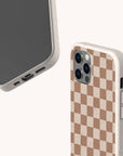 Two smartphones with beige speckled cases; one displays the screen, while the other shows the back featuring the Almond Checker Eco-Friendly Phone Case in a brown and beige checkerboard pattern. These eco-friendly cases include precise camera and port cutouts, combining classic style with practical convenience.