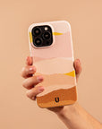 A hand holds the Mountains Eco-Friendly Phone Case, featuring an abstract, layered design in pink, peach, and yellow on beige. Compatible with iPhone and Samsung Galaxy models.