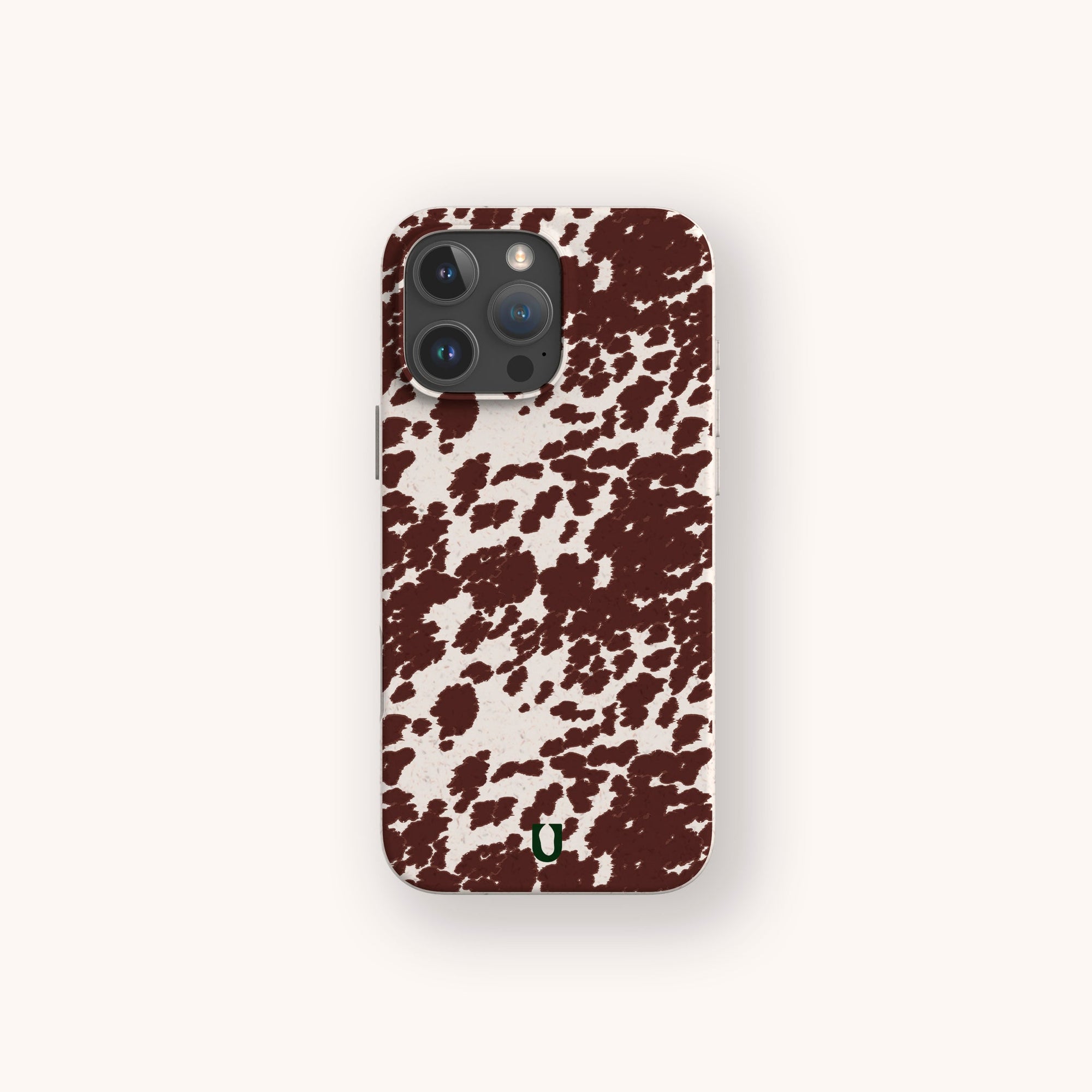 The Brown Cow Print Eco-Friendly Phone Case, compatible with iPhone and Samsung Galaxy, features irregular brown splotches on white and a small green horseshoe near the bottom, shown against a plain light background.