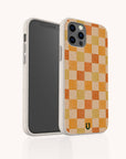 An iPhone or Samsung Galaxy in a Brown and Orange Checker Eco-Friendly Phone Case stands upright, screen facing left, displaying the patterned back with a small green and yellow logo at the bottom.