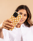 A woman in a white shirt takes a selfie against a beige background, holding up her smartphone fitted with the Star Terracotta Checker Eco-Friendly Phone Case featuring a yellow and white checkered pattern.