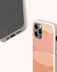 Two smartphones in speckled beige Mountains Eco-Friendly Phone Cases; one displays the screen and bottom ports, while the other shows the camera and a pink, peach, and orange abstract wave design on the back. These eco-friendly Mountains cases are compatible with iPhone and Samsung devices. Background is off-white.