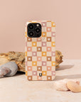 The Star Pink Checker Eco-Friendly Phone Case, made from plant-based materials in star pink checker, beige, brown, and yellow, sits upright on a stone surface with rocks and a light beige background behind it. Stylish and sustainable.