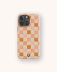 The Pink Watercolour Checker Eco-Friendly Phone Case features a peach, pink, and beige checkerboard design with a small black horseshoe-shaped logo at the bottom center on an off-white background. Compatible with iPhone and Samsung Galaxy.