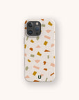The Mini Confetti Eco-Friendly Phone Case features a beige terrazzo-style design with abstract green, yellow, orange, and pink shapes. Made from compostable materials, it fits iPhone and Samsung Galaxy models and has a small dark green "U" logo at the bottom center.