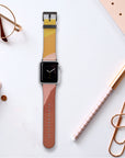 On a desk surrounded by a planner, rose gold pen, glasses, and golden paperclip lies an Apple Watch featuring the Abstract Cut Out II Vegan Leather Band with its trendy, multicolored strap. The watch face displays an analog clock, and its cruelty-free faux leather band beautifully complements the desk's soft, neutral aesthetic.