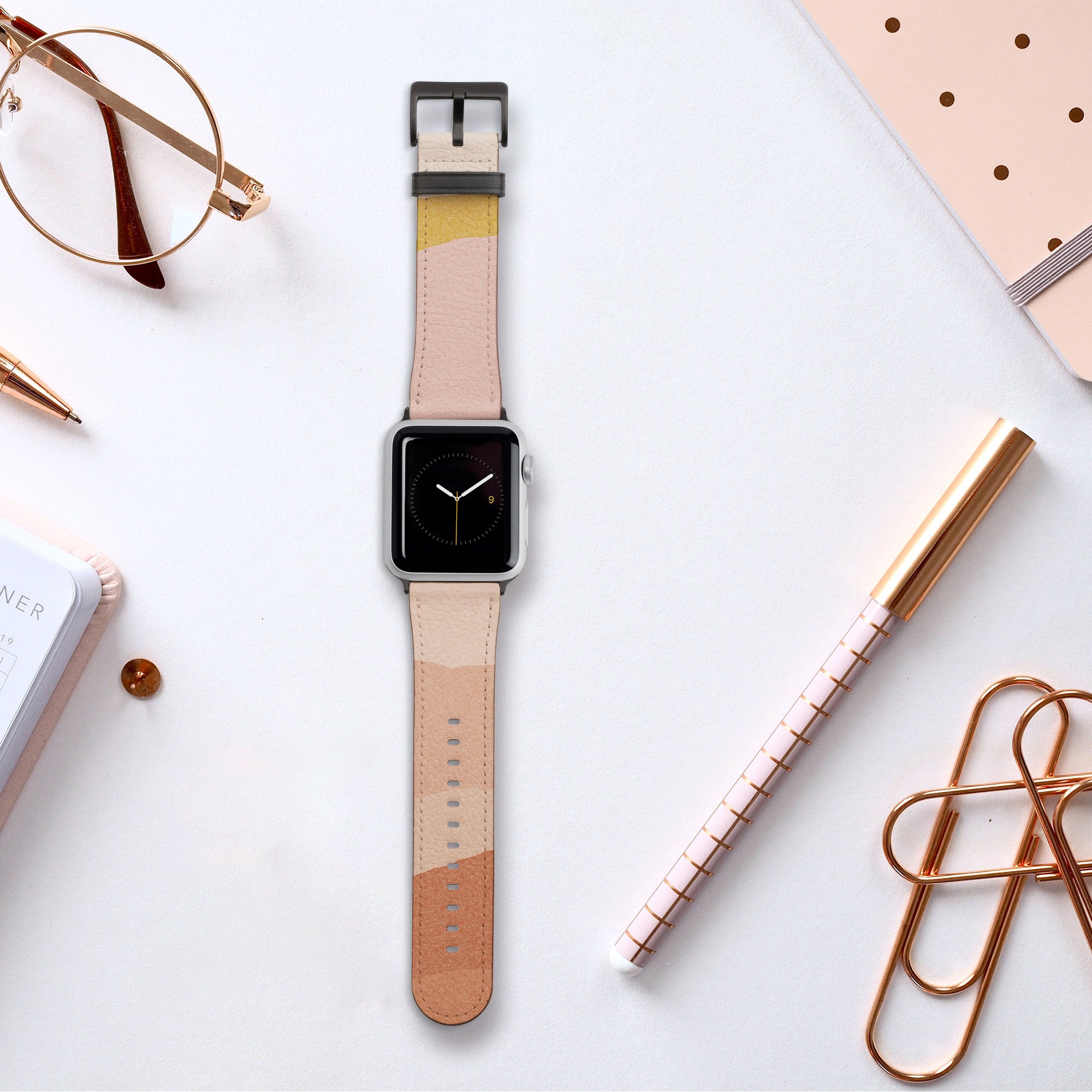 An Apple Watch adorned with the Abstract Cut Out III Vegan Leather band, featuring a beige and peach cruelty-free design, rests on a white surface. It is accompanied by a notebook, a grid-patterned pen, glasses, a binder clip, and polka-dotted paper. The watch face shows a minimalist clock against an obsidian backdrop.