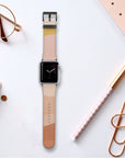 An Apple Watch adorned with the Abstract Cut Out III Vegan Leather band, featuring a beige and peach cruelty-free design, rests on a white surface. It is accompanied by a notebook, a grid-patterned pen, glasses, a binder clip, and polka-dotted paper. The watch face shows a minimalist clock against an obsidian backdrop.