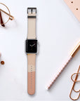 A smartwatch with a Valleys Vegan Leather Apple Watch Band in two-toned beige and brown rests on a white surface, surrounded by rose gold paper clips, a checkered pen, a notebook, round glasses, and an eco-conscious planner.