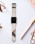The Beige Abstract Lines Vegan Leather Apple Watch Band, featuring a beige and gold-patterned strap, is elegantly displayed on a white surface. Surrounding it are rose gold stationery items like glasses, binder clips, a pen, and a dotted notebook.