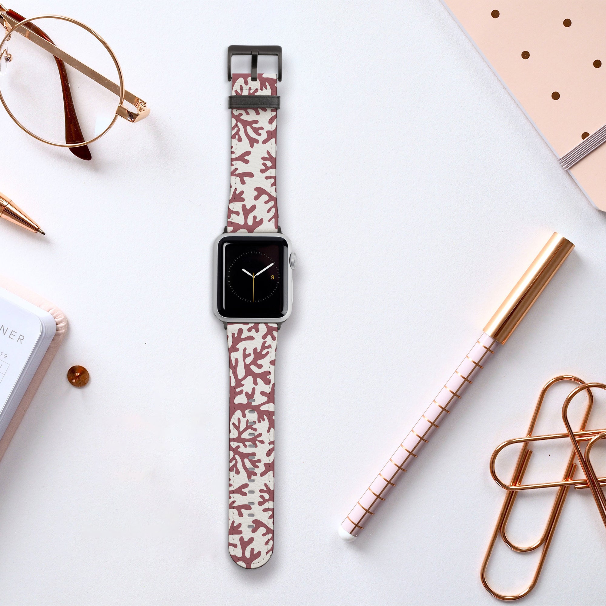A smartwatch featuring the Brown Coral Vegan Leather Apple Watch Band, adorned with a decorative pink and white strap made from eco-friendly vegan leather, rests on a flat surface. It's surrounded by office supplies such as a notebook, pens, paper clips, and glasses—ideal for the conscious consumer.