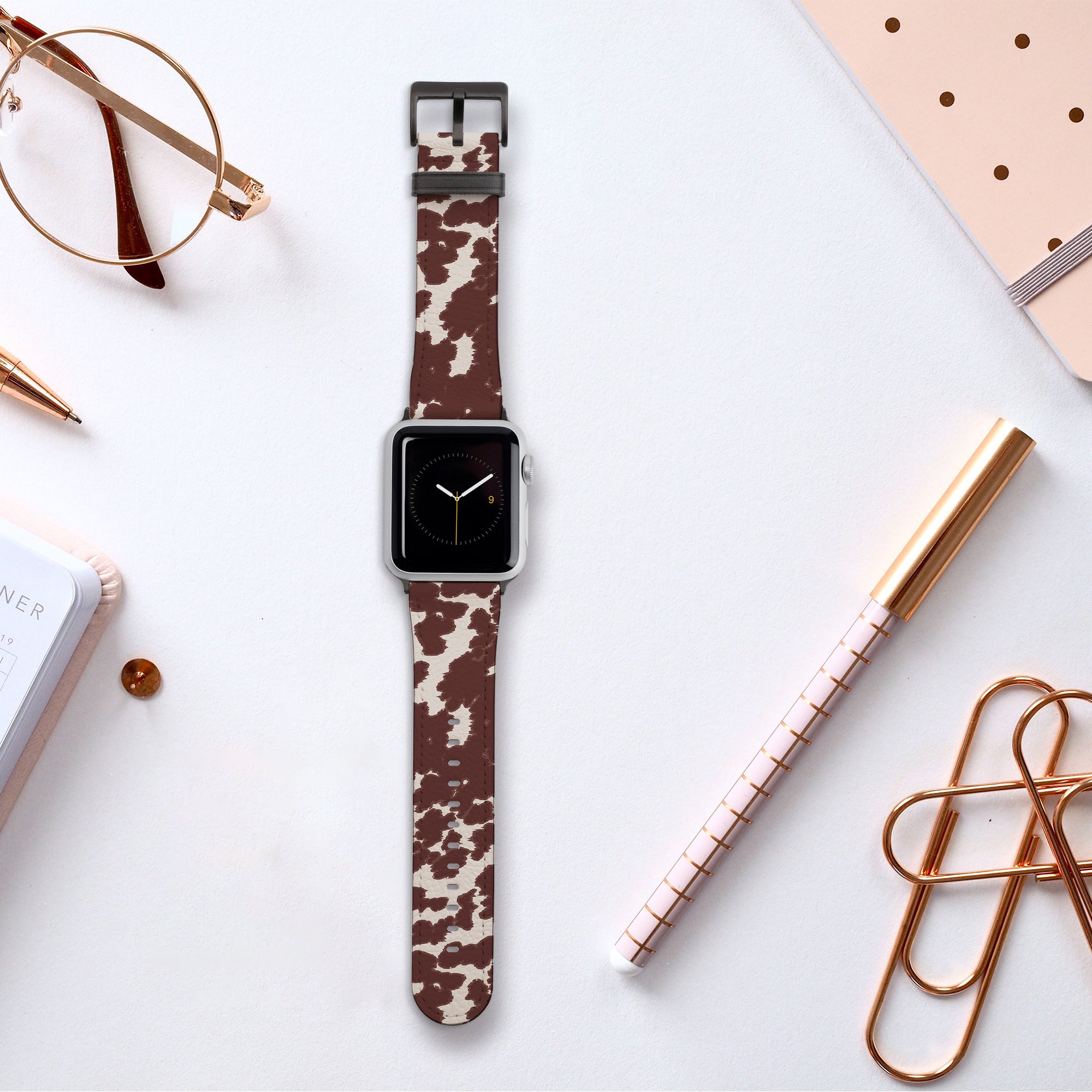 A Brown Cow Vegan Leather Apple Watch Band sits atop a white surface, surrounded by rose gold and white office supplies such as a notebook, pen, paper clips, and glasses.