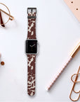 A Brown Cow Vegan Leather Apple Watch Band sits atop a white surface, surrounded by rose gold and white office supplies such as a notebook, pen, paper clips, and glasses.