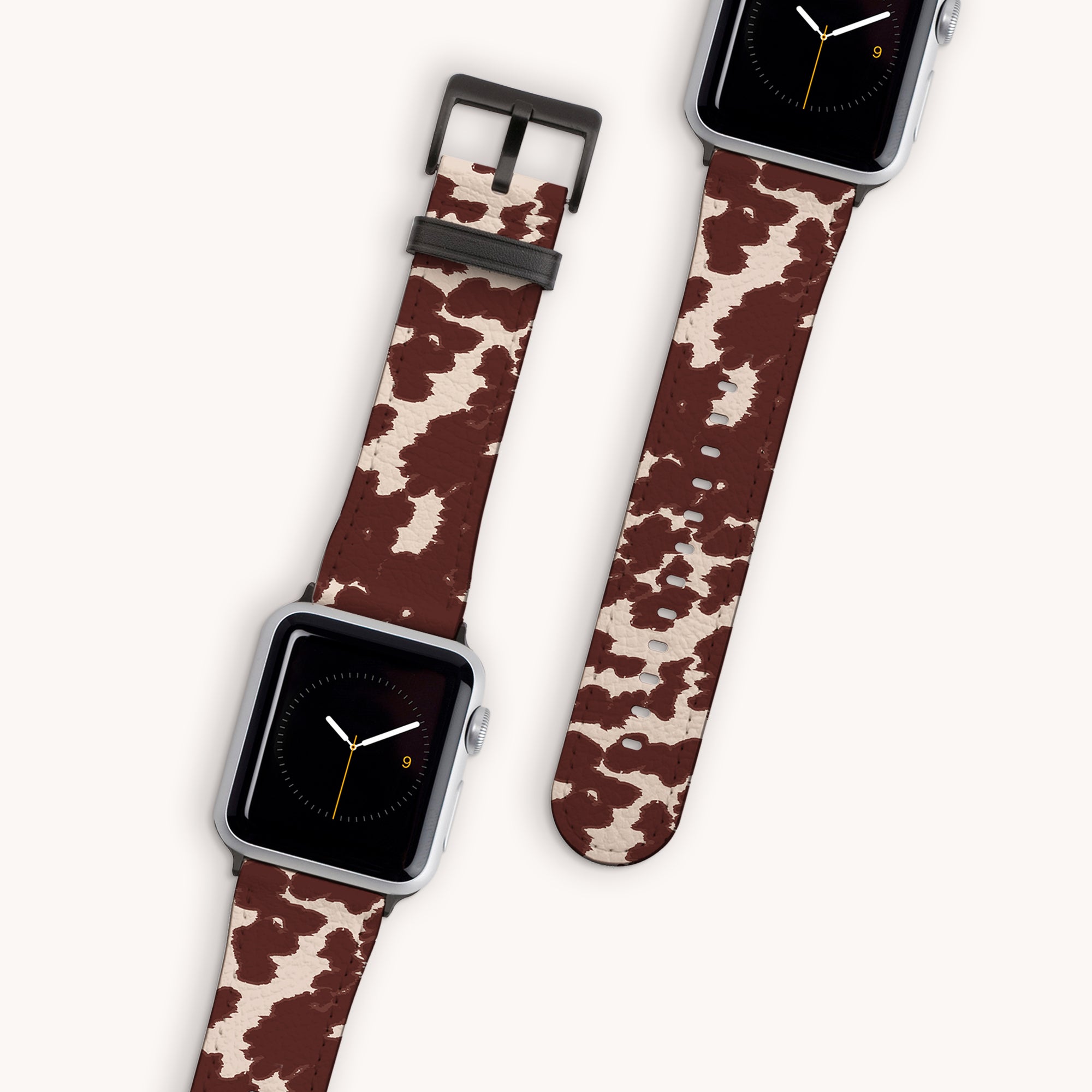 Displayed on a plain background are two smartwatches featuring the Brown Cow Vegan Leather Apple Watch Band, showcasing a rustic charm. The watch faces depict an analog clock with a minimalist design against a black backdrop, while the bands highlight a distinct brown and white cowhide pattern.