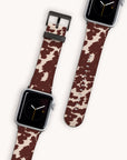 Displayed on a plain background are two smartwatches featuring the Brown Cow Vegan Leather Apple Watch Band, showcasing a rustic charm. The watch faces depict an analog clock with a minimalist design against a black backdrop, while the bands highlight a distinct brown and white cowhide pattern.