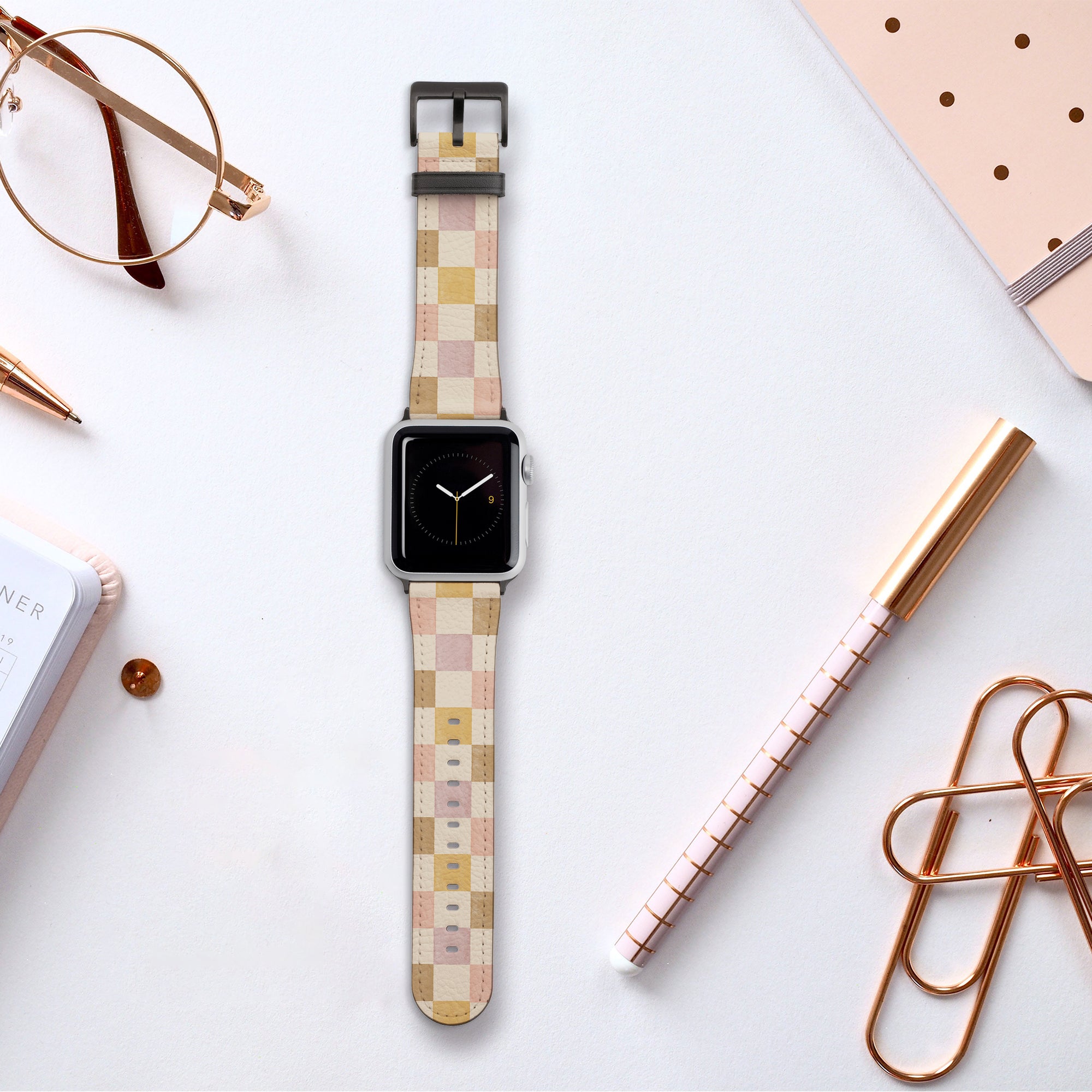 A smartwatch featuring a Brown and Purple Checker Vegan Leather Apple Watch Band rests on a white surface. It's surrounded by a notebook, a pen, glasses, and an additional pen with gold accents. The arrangement is minimalistic and organized, highlighting sustainable craftsmanship.