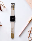 A smart watch featuring the Caramel Abstract Strokes Vegan Leather Apple Watch Band in beige and gold rests on a desk. It is surrounded by a gold pen, a pair of glasses, a planner, a notebook, and large gold paper clips, contributing to the desk's modern and organized aesthetic.