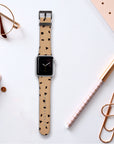 On a desk, the Geo Shapes in Brown Vegan Leather Apple Watch Band rests elegantly, featuring a geometric-patterned beige strap. Surrounding it, minimalistic items such as a notebook, paper clips, a pen, glasses, and a pencil in gold and neutral shades create an effortlessly chic ambiance.