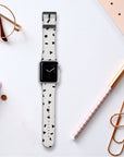 A smartwatch adorned with the "Geo Shapes in White" vegan leather Apple Watch band rests on a white surface, surrounded by a notebook, rose gold paper clips, a striped design pen, glasses, and an elegant white band featuring black geometric patterns.