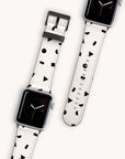 Two Apple Watches adorned with the "Geo Shapes in White Vegan Leather" bands are showcased with a pattern of black geometric shapes on white vegan leather. The watch faces exhibit the time through an analog-style clock, and the watches are set against a light, neutral background.