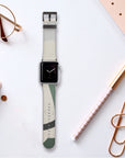 The Green Abstract Lines Vegan Leather Apple Watch Band on a white surface, surrounded by a notebook, glasses, a pen, and gold paperclips. The watch face shows an analog clock display.