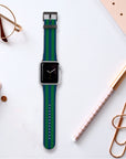 A smartwatch featuring a Green Blue Stripes Vegan Leather Apple Watch Band is placed on a desk next to rose gold glasses, a notebook, a pen, a pencil, and oversized paper clips.