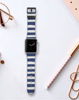 The Navy Stripes Vegan Leather Apple Watch Band rests on a white surface, surrounded by stationery items like a pen, notebook, paperclips, and reading glasses. The watch display elegantly shows an analog clock face.