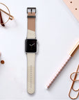 A smartwatch adorned with the Neutral Abstract Vegan Leather Apple Watch Band, showcasing a beige and brown strap, is set on a white surface amid a notebook, pen, paper clips, glasses, and a planner. The watch screen displays a simple clock face.