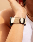 A close-up showcases a person wearing the Neutral Abstract Vegan Leather Apple Watch Band, featuring beige vegan leather with black accents. As they gently touch their face, they're complemented by a white sleeveless top and a gold chain necklace, achieving an elegant yet modern look.
