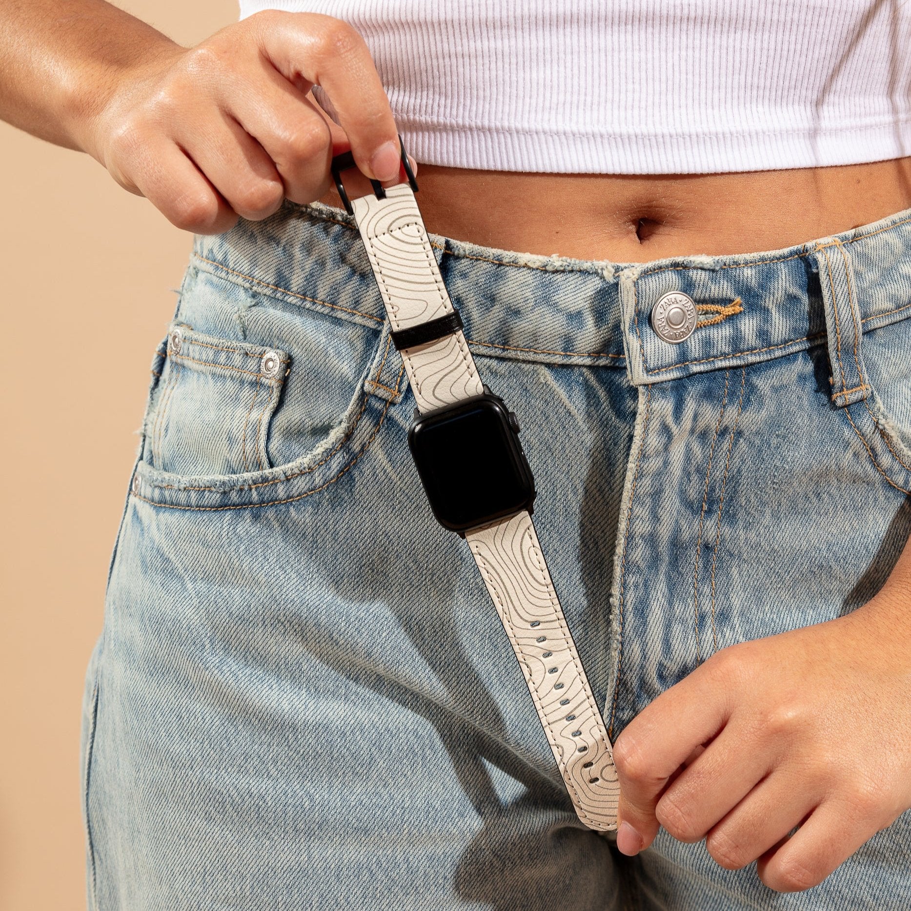 A person in a white crop top and blue jeans is adjusting the Terrain Vegan Leather Apple Watch Band, crafted from eco-friendly materials, looping it around a belt loop. The black watch face complements the jeans' button with intricate stitching details.