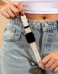 A person in a white crop top and blue jeans is adjusting the Terrain Vegan Leather Apple Watch Band, crafted from eco-friendly materials, looping it around a belt loop. The black watch face complements the jeans' button with intricate stitching details.