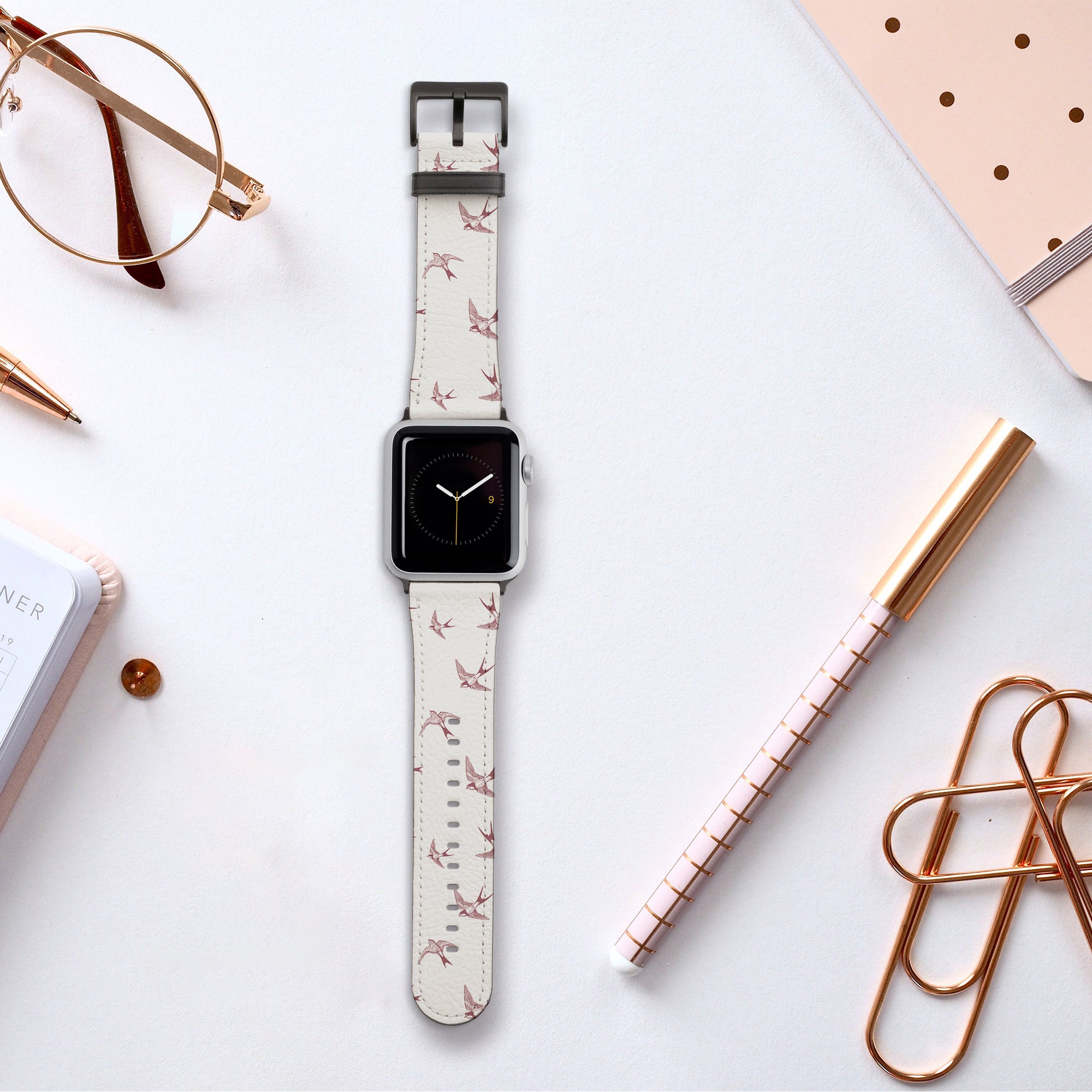 The Swallows Vegan Leather Apple Watch Band with its bird-themed design rests on a white desk, surrounded by a notebook, golden paperclips, a pen, and eyeglasses, creating an elegant workspace setup.