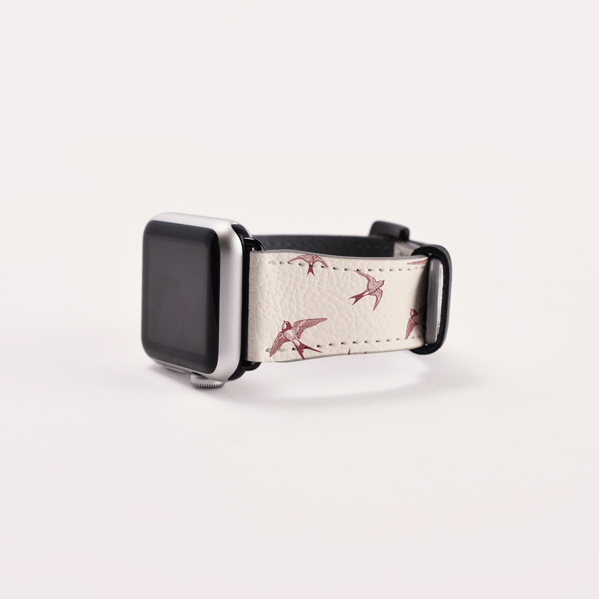 The Swallows Vegan Leather Apple Watch Band features an eco-friendly, bird-themed strap adorned with small red birds on a white band. The black watch face is off, highlighting its decorative design against a plain white background.
