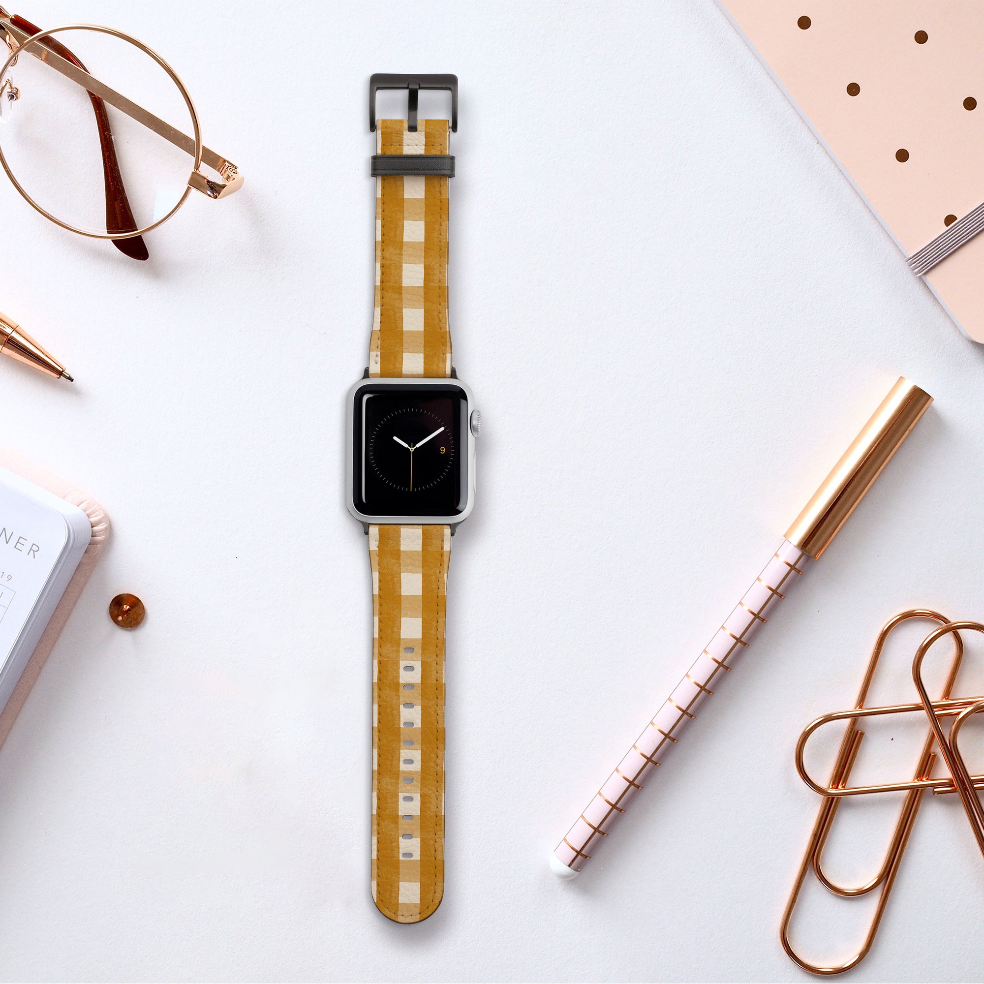 A Terracotta Gingham Vegan Leather Apple Watch Band lies on a white desk surrounded by rose gold paperclips, a notebook, glasses, pens, and a dotted beige planner.