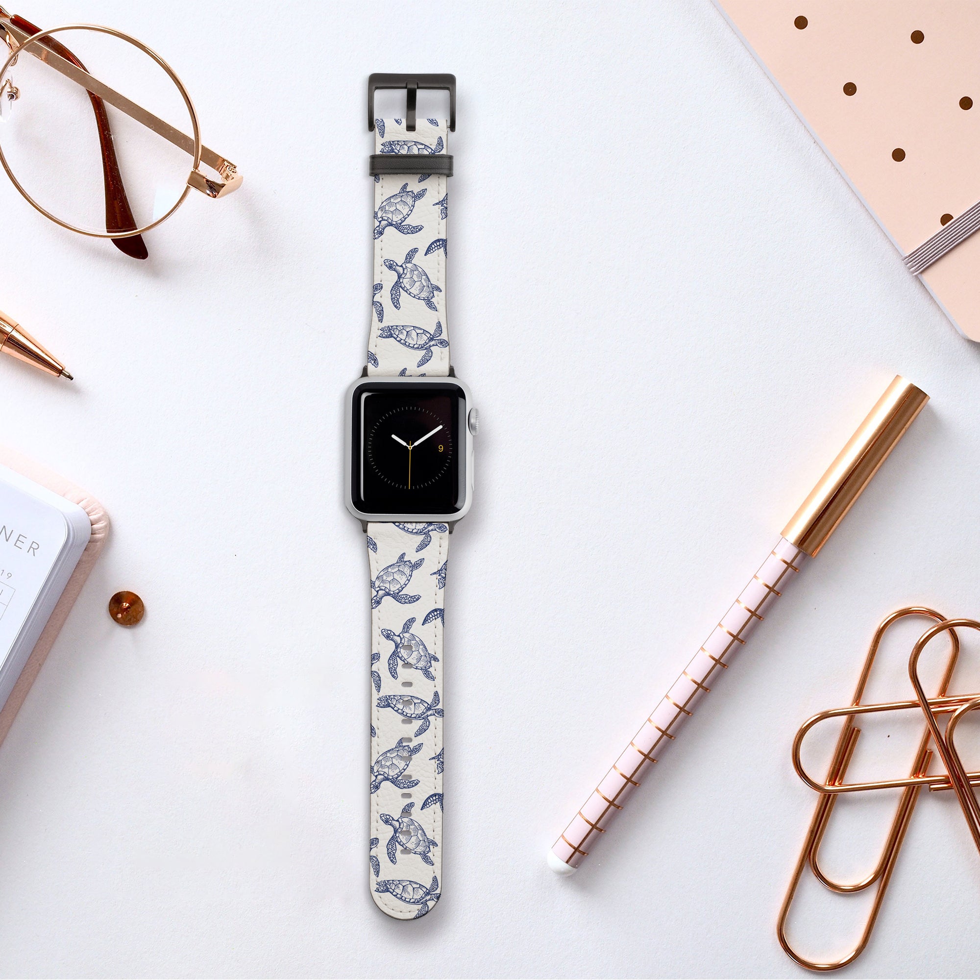 On a desk lies a smartwatch donned with the Turtles Vegan Leather Apple Watch Band, showcasing a white strap and blue bird pattern, accompanied by a notebook, rose gold pen, glasses, and large paperclips.