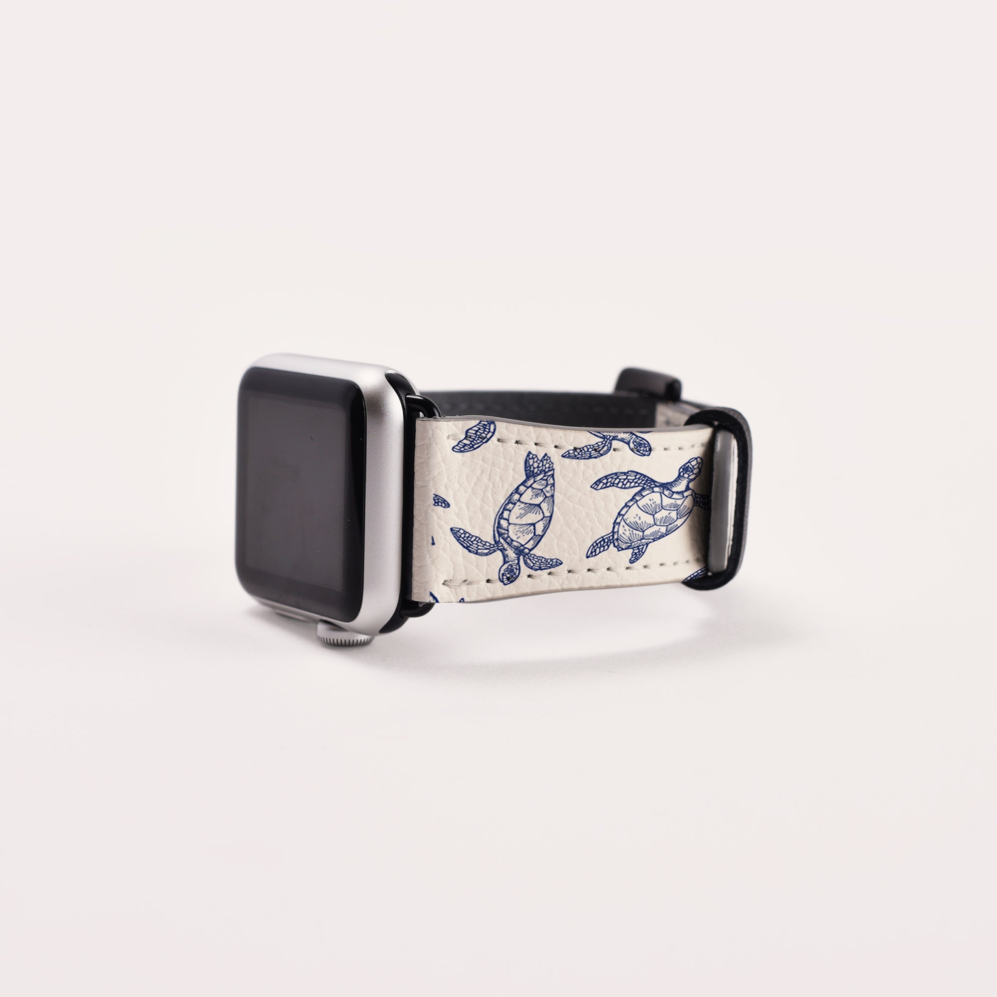 The Turtles Vegan Leather Apple Watch Band is an eco-friendly smartwatch strap in white and blue with a turtle design. It features a black watch face and partially looped strap set against a plain white background.