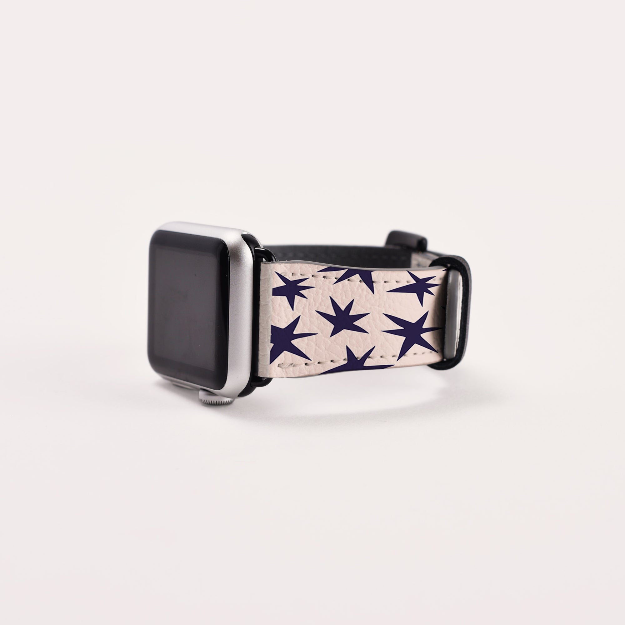 The Wonky Stars Vegan Leather Apple Watch Band features dark blue stars on a white background. Its eco-conscious design adds celestial charm to your wrist while resting gracefully on a light surface.