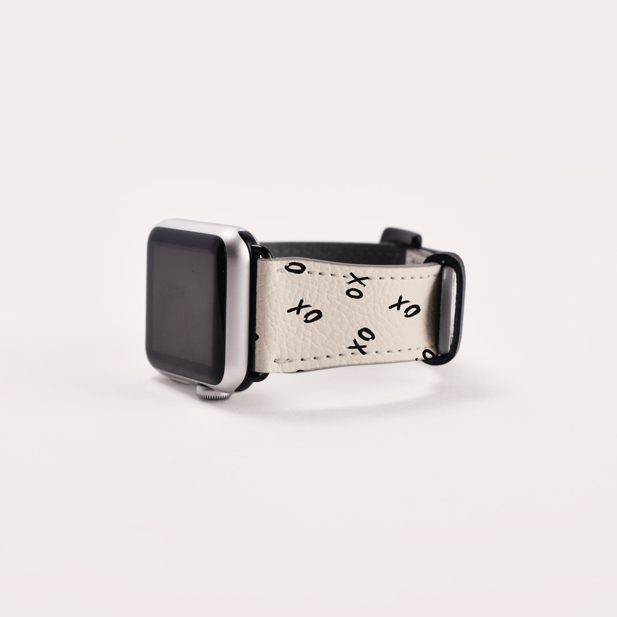 A smartwatch with a silver face features the Xo Xo Vegan Leather Apple Watch Band in beige, adorned with black "XO" symbols and black hardware. This eco-conscious band is elegantly angled against a plain white background.