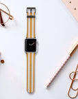 An Apple Watch featuring the Orange Stripes Vegan Leather Band lies on a desk alongside a notebook, gold pen, glasses, large paper clip, and closed laptop. The watch screen shows an analog clock face.