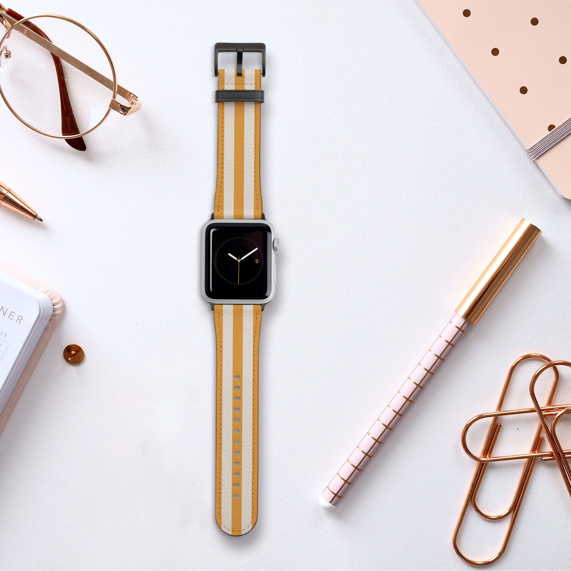 A Yellow Beige Stripes Vegan Leather Apple Watch Band with water resistance sits on a white desk amidst office supplies, including a gold pen, notebook, paperclips, and glasses.