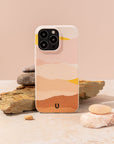 The Mountains Eco-Friendly Phone Case, featuring a pastel abstract mountain design, stands upright on a rough stone amid smooth pebbles against a light beige background.