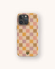 A smartphone with a camera bump is protected by the Pink and Purple Checker Eco-Friendly Phone Case, featuring a checkered pattern in beige, pink, tan, and brown squares. A small green horseshoe-shaped logo appears at the bottom center of the case.