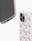 Two smartphones with beige cases are shown. One features the “Pink Coquette Flowers Eco-Friendly Biodegradable Phone Case,” showcasing a vintage floral design adorned with pink and blue blossoms, while the other case is plain. Both phones display the camera, side, and charging port area against a light background.