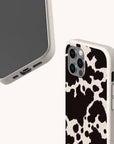 Two smartphones in protective cases; one features the Cow Print Eco-Friendly Phone Case in bold black and white, while the other is a plain beige eco-friendly case—both compatible with iPhone and Samsung devices—displayed on a plain, light background.