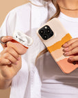 A person holds an open AirPods case in one hand and a smartphone with a colorful Mountains Eco-Friendly Phone Case in the other, while wearing a white shirt, gold necklace, and rings.